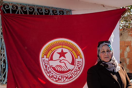 //Le operaie davanti alla Mamotex chiusa, afferrano uno striscione che rivendica il loro diritto al lavoro, sopra di loro la bandiera dell’UGTT (l’Union Générale des Travailleurs Tunisiens), il potente sindacato dei lavoratori tunisini. Foto di Monia Ben Hamadi.