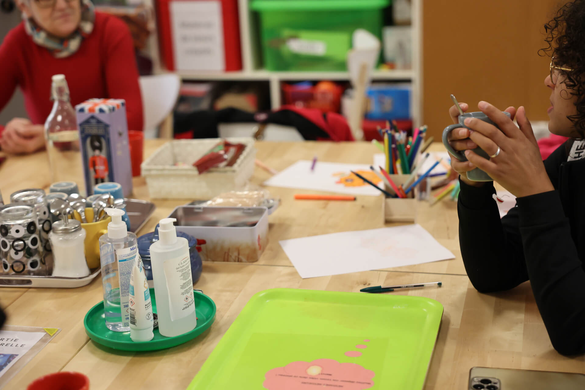 Scène de vie et de discussion entre plusieurs personnes. Sur la table, on peut voir divers feuilles et crayons, et un plateau avec des tasses de thé. | CESS-IDF - Collectif Epiceries IDF